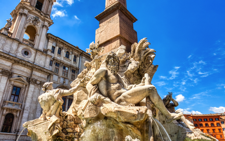 Brunnen mit Skulpturen und Obelisk auf einem Platz vor einem historischen Gebäude
