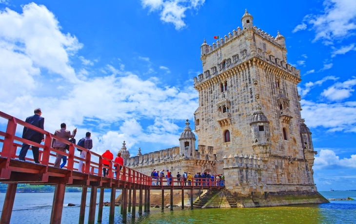 Menschen auf Brücke vor dem Torre de Belém in Lissabon bei blauem Himmel.