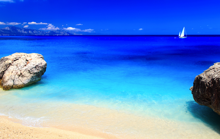Strand mit türkisblauem Wasser, Felsen und Segelboot am Horizont.
