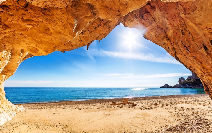 Blick aus Höhle auf sonnigen Sandstrand und blaues Meer.