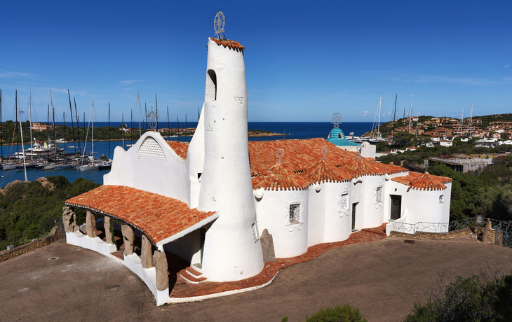 Weiße Kirche mit rotem Dach nahe einem Yachthafen, blauer Himmel im Hintergrund.