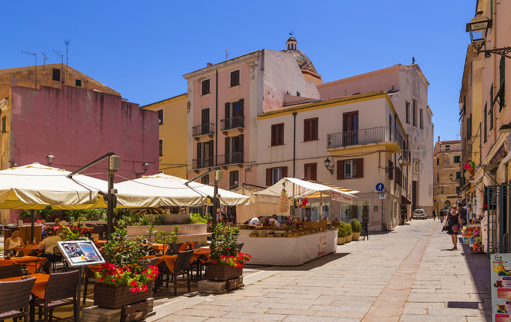 Straßenszene in einer sonnigen mediterranen Stadt mit Cafés und bunten Gebäuden.