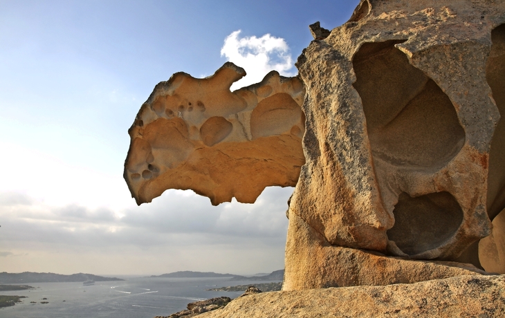 Felsformation am Meer mit blauem Himmel und Wolken im Hintergrund.