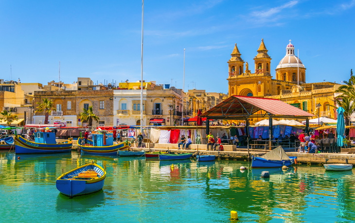 Bunte Fischerboote im Hafen vor historischen Gebäuden und einem Markt in Malta.