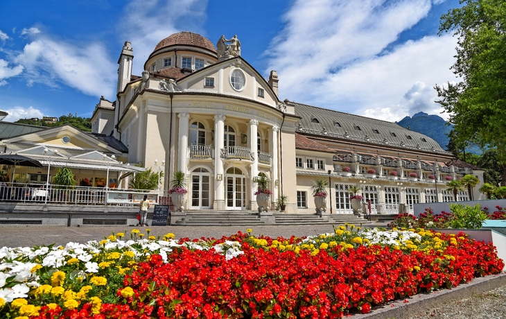 Historisches Gebäude mit Blumenbeet im Vordergrund und Bergen im Hintergrund.
