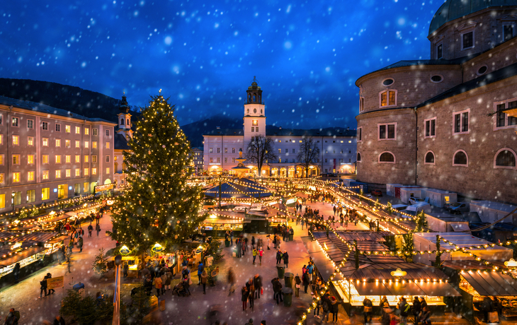 Weihnachtsmarkt bei Nacht mit erleuchtetem Baum und schneebedecktem Himmel.