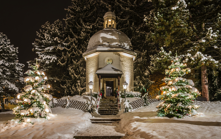Schneebedeckte Kapelle mit beleuchteten Weihnachtsbäumen in winterlicher Nacht.