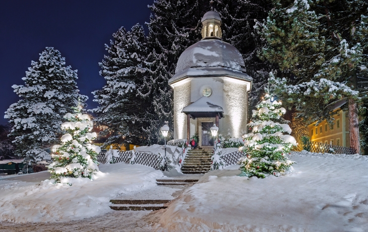 Verschneite Kapelle bei Nacht mit beleuchteten Weihnachtsbäumen.