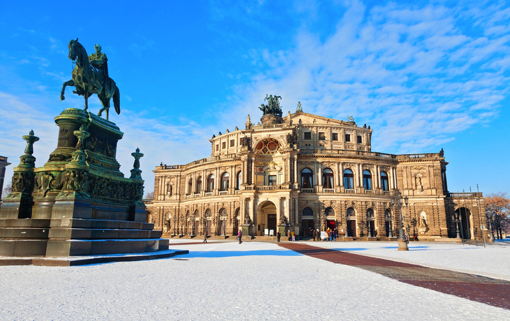 Dresdner Semperoper im Winter mit Schnee auf dem Theaterplatz und einer Statue im Vordergrund.