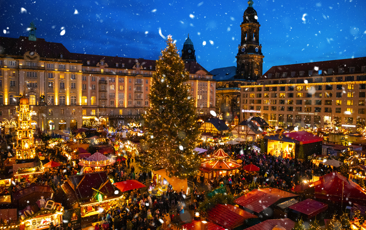 Weihnachtsmarkt am Abend mit Lichtern und großem geschmücktem Tannenbaum im Schnee.
