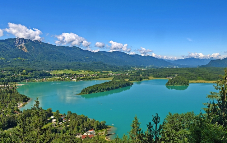 Panoramabild von einem türkisfarbenen See mit bewaldeten Ufern und Bergen im Hintergrund.
