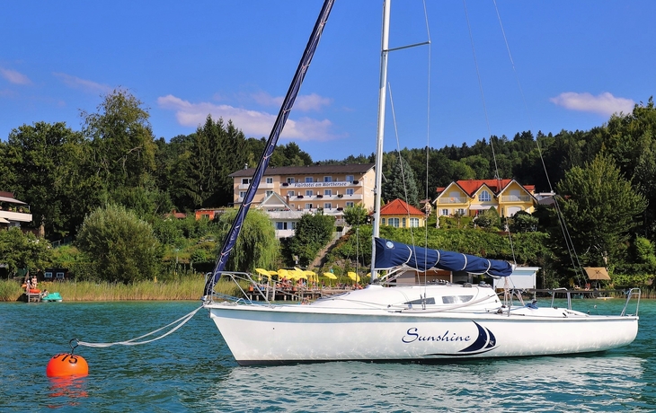 Segelboot auf einem See vor einer bewaldeten Küstenlandschaft.