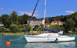 Segelboot auf einem See vor einer bewaldeten Küstenlandschaft.