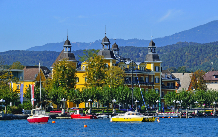 Gelbes Gebäude mit Türmen am See, Boote im Vordergrund, Berge im Hintergrund.