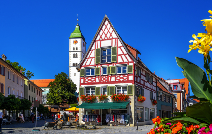Fachwerkhaus mit Uhrturm im Hintergrund, blauer Himmel, bunte Blumen im Vordergrund.