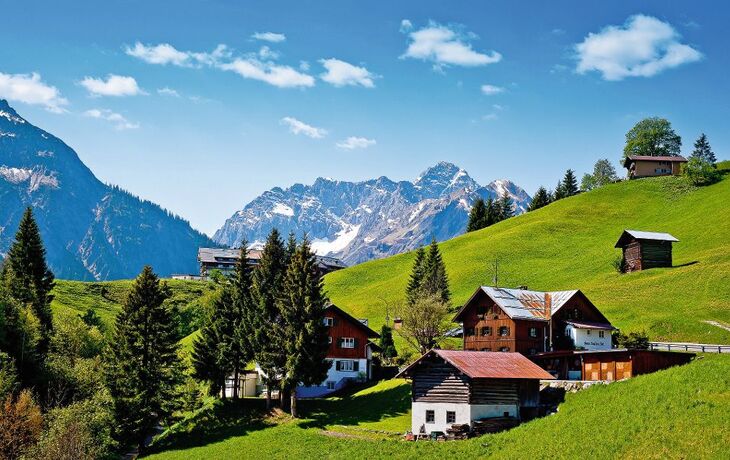Dorf in den Alpen mit Holzhäusern, grünen Wiesen und Bergen im Hintergrund
