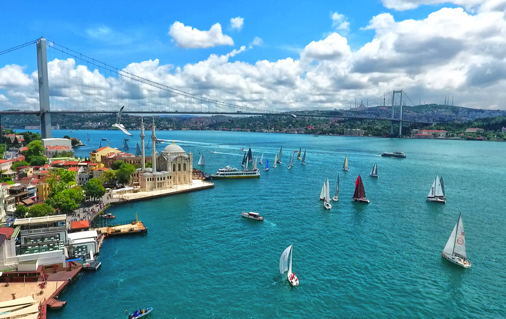 Blick auf die Bosporus-Brücke in Istanbul mit Segelbooten im Vordergrund und einer Moschee am Ufer.