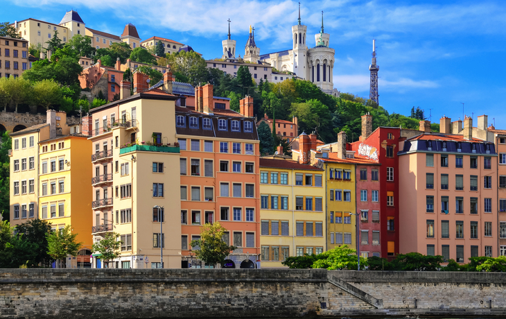 Bunte Gebäude am Flussufer mit Hügel und Kirche im Hintergrund.